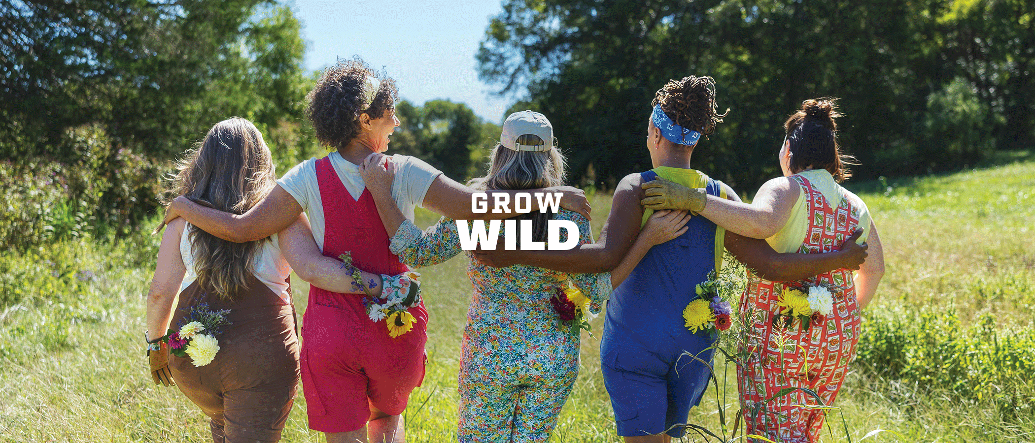 Grow Wild. Women in colorful overalls linking arms and walking through a field