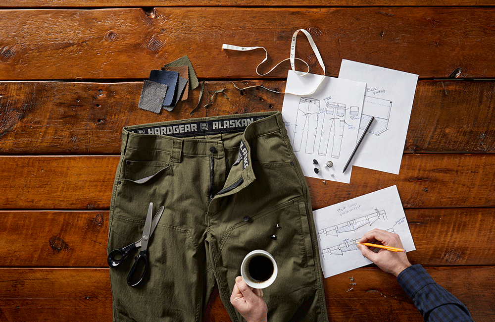 Olive Stone Run pants laid out on workshop bench with paper and pens